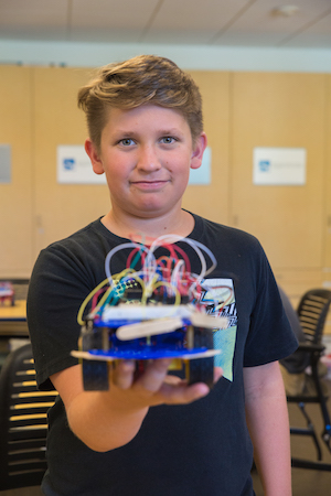 A middle school student looks into the camera. His hand holds a small robotic device. He holds this robotic device in front of him, arm outstretched toward the camera.