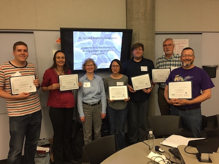 Seven people stand in front of a television screen in a classroom. Six of the seven are holding certificates of commendation noting their participation in the HHMI funded Northstar Summer Institute.